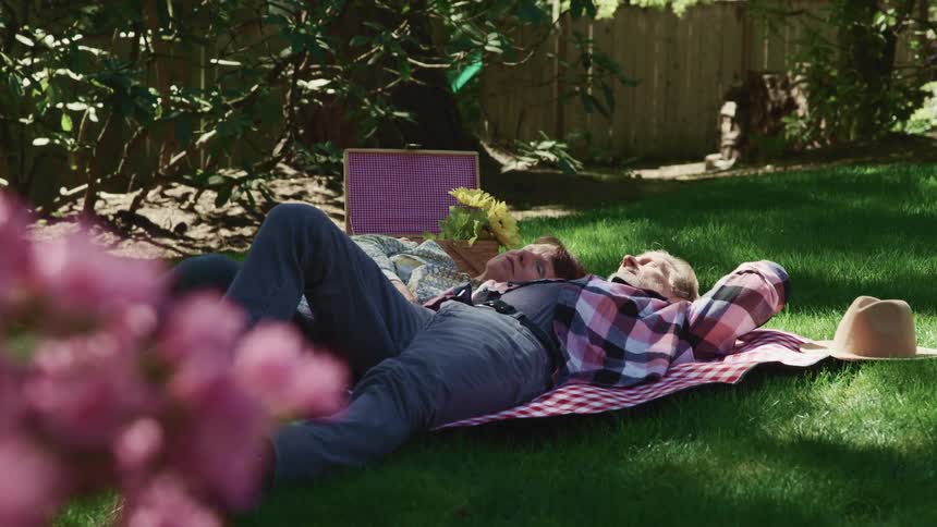 A man and woman sleep side by side on a picnic blanket in a fenced off yard