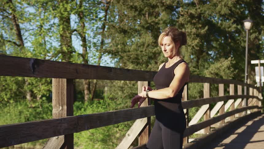 Woman in sport clothes checks her wristwatch while standing in a wooden bridge near the forest