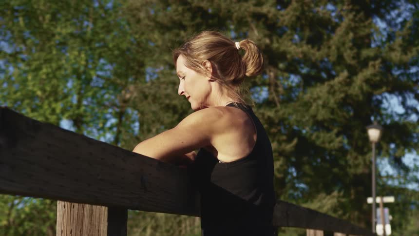 Woman leans against wooden rail looking down near trees
