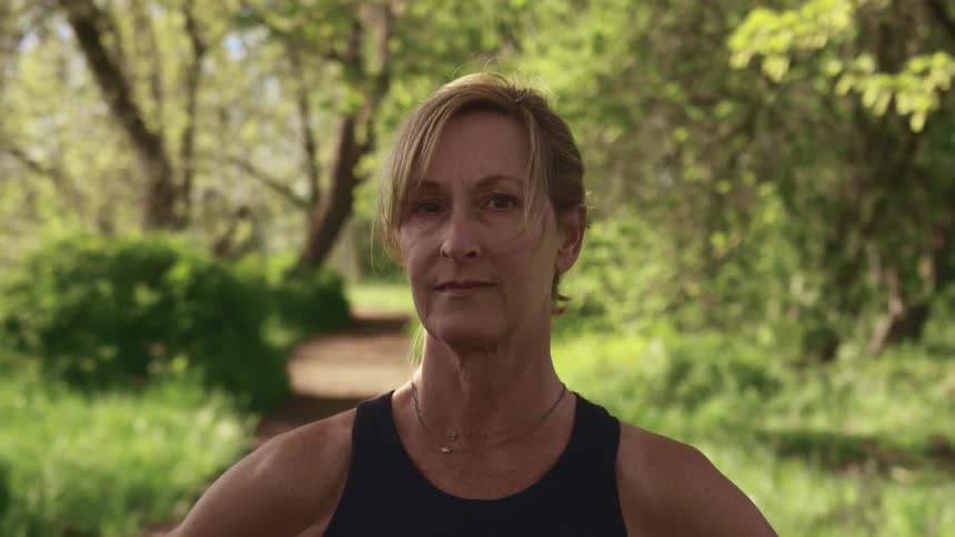Elderly woman with a ponytail stands on a dirt road and faces the camera with eye contact in the park