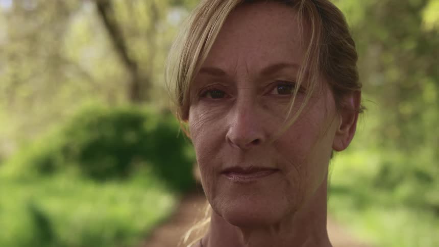 Blonde woman with wrinkles standing on a dirt road faces the camera with eye contact in a forest