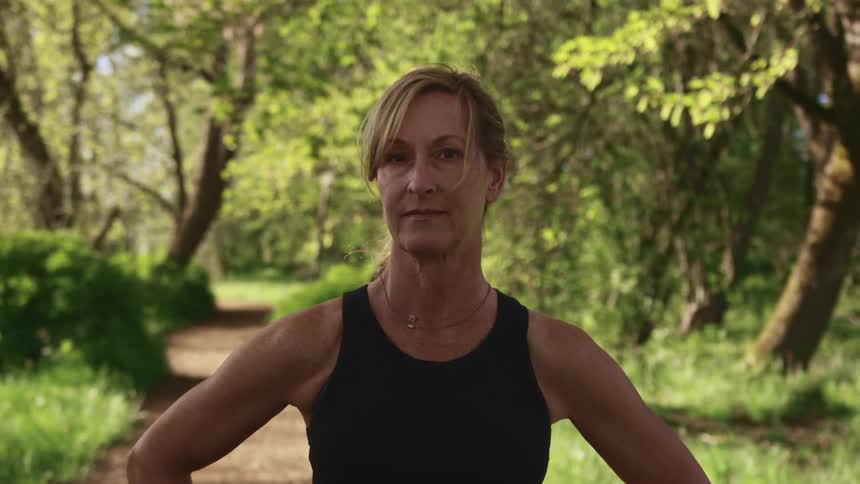 Middle aged blonde woman with a ponytail stands on a dirt road and faces the camera with eye contact in a park