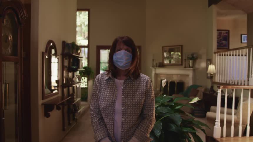 Middle aged woman with brown hair and a light blue mask raises her head and faces the camera with eye contact in the living room at home