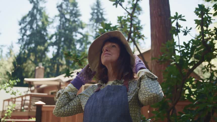 An elderly woman stands outside her home with her gardening gloves on and looks up at the sky