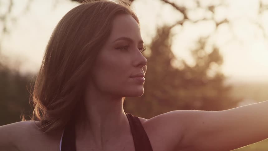 Woman with brown hair and makeup spreads her arms wide in an open field at sunset