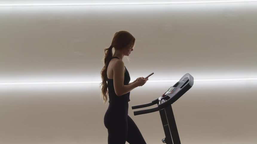 A female teenager walks on a treadmill while looking at her phone