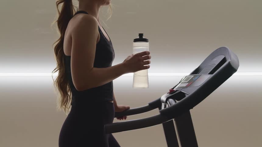 A woman stands motionlessly on a treadmill holding a bottle containing gently vibrating water
