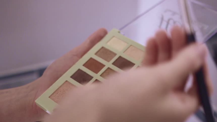 A womans hand holding a makeup brush and makeup dipping the brush into makeup and raising it up
