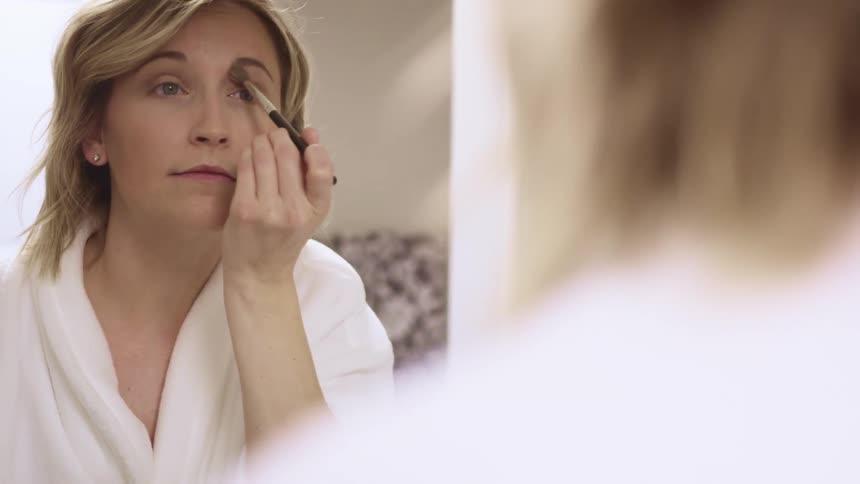 Female wearing bathrobe looks in the mirror as she applies eye shadow using a soft makeup brush