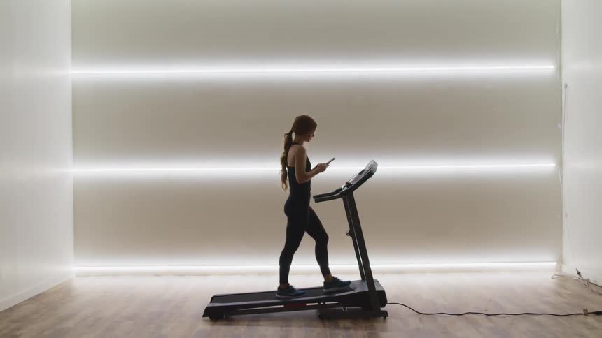 Woman with a ponytail tights and running shoes walks on a treadmill while using a smartphone in a room