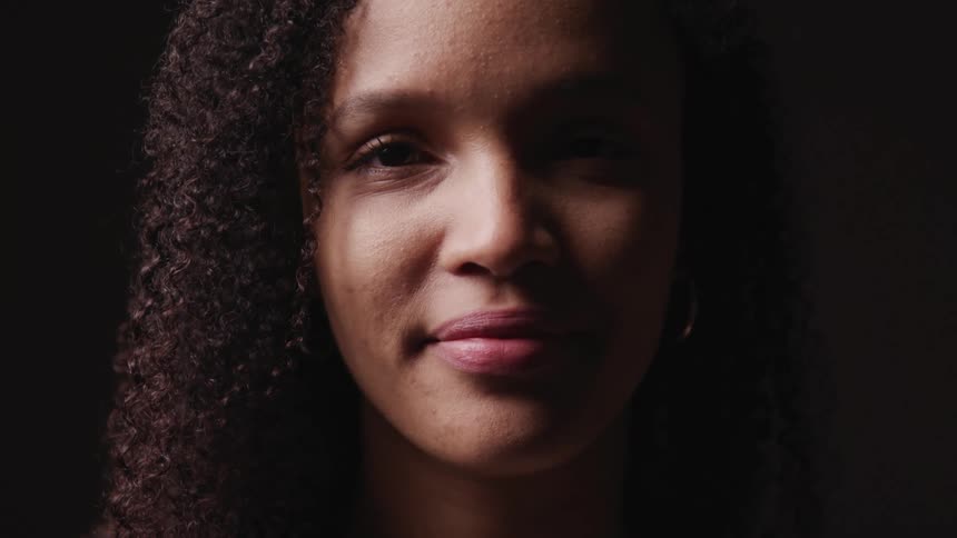 Black woman with curly hair and earrings faces the camera with eye contact on a dark background