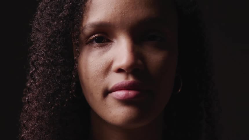 Black woman with curly hair and earring raises her head and faces the camera with eye contact on a dark background