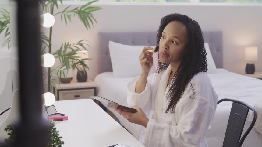 Woman holds a cosmetic tray and applies makeup to her face with a brush while sitting in a bedroom