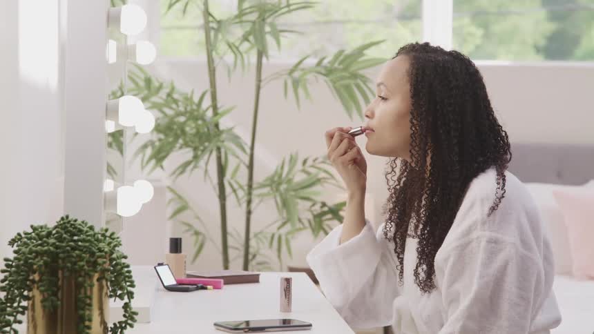 Woman with curly hair and a bathrobe looks in the mirror and applies lipstick in her room