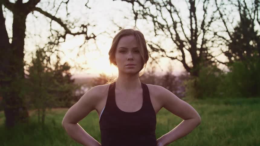 Woman wearing a tank top stands in a field near the trees with her hands on her hips