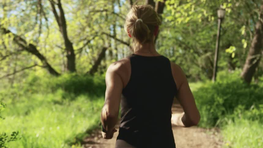 A woman in athletic wear jogs along a dirt path through a woodland on a sunny day