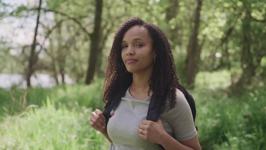 Woman with curly hair a grey t shirt and a black backpack stands and looks at the camera in the forest