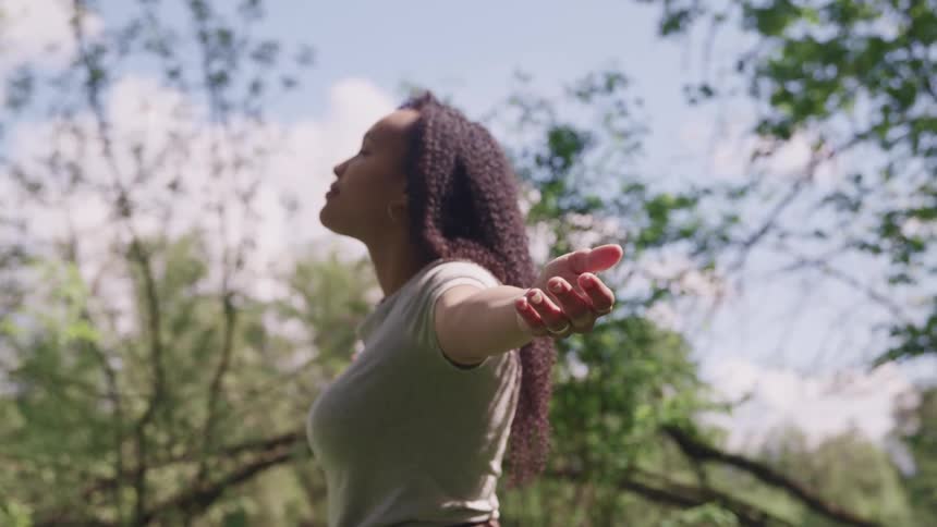 A woman stands in a forest with her eyes closed and arms raised up to her sides