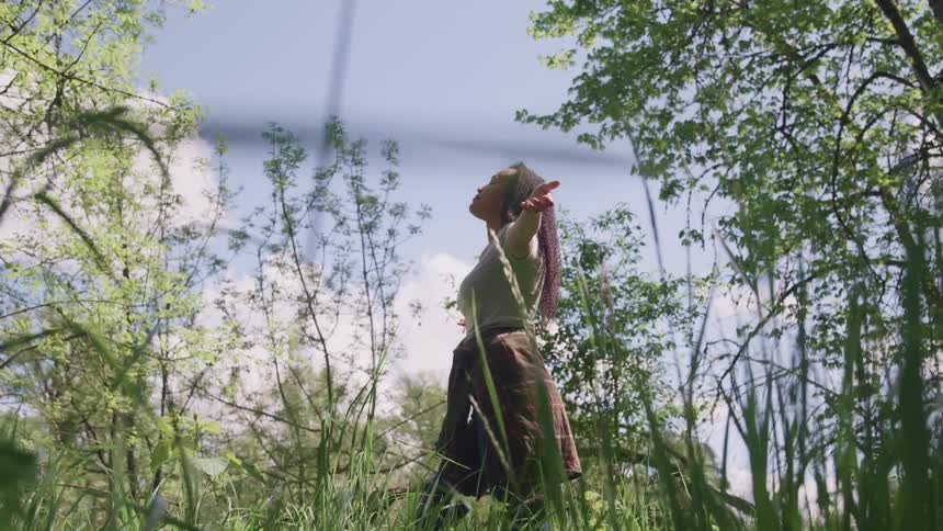 A woman stands in a field of tall grass near a forest and raises her arms up to her sides