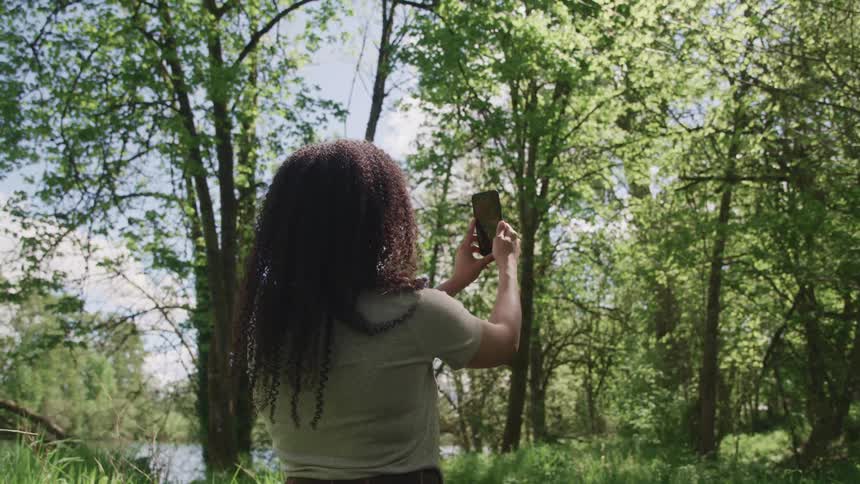 Black woman with curly hair and a grey t shirt takes a picture of tall trees with a black smartphone in the forest