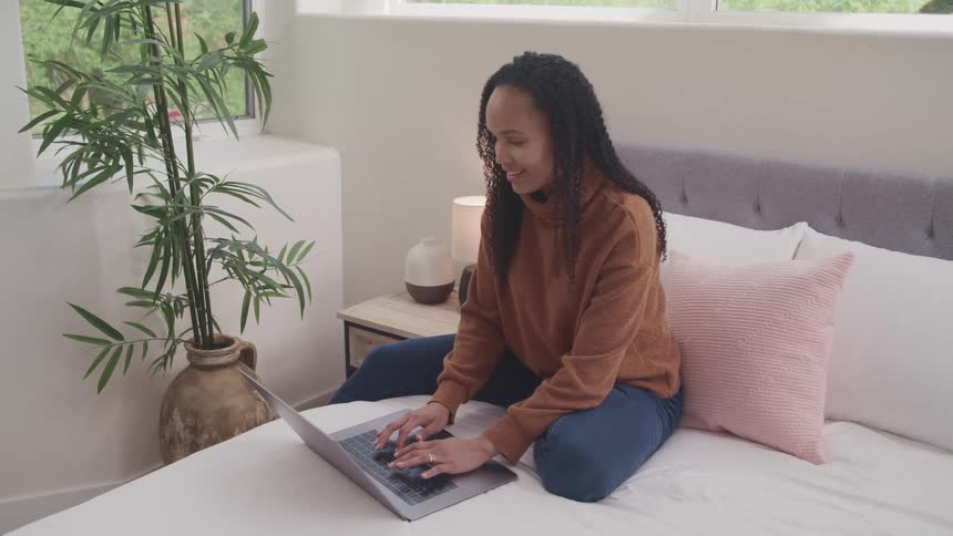 Woman sits on bed typing on laptop and smiling
