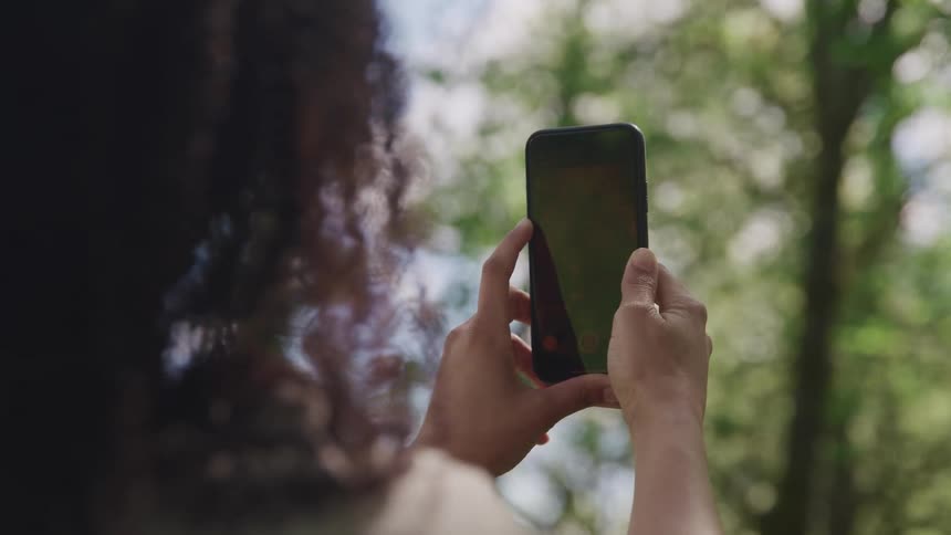 Black woman with curly hair takes a picture of tall trees with a black smartphone in the forest