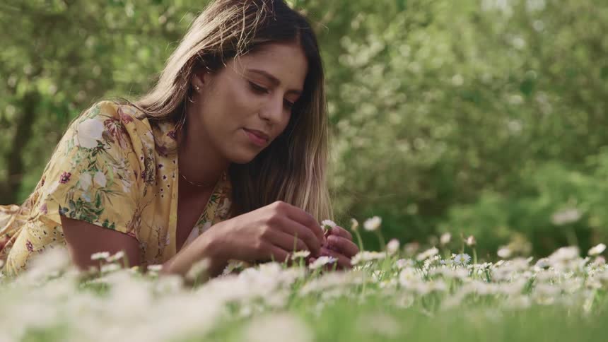 A teenage girl lying in a field looks at a flower and picks the petals off