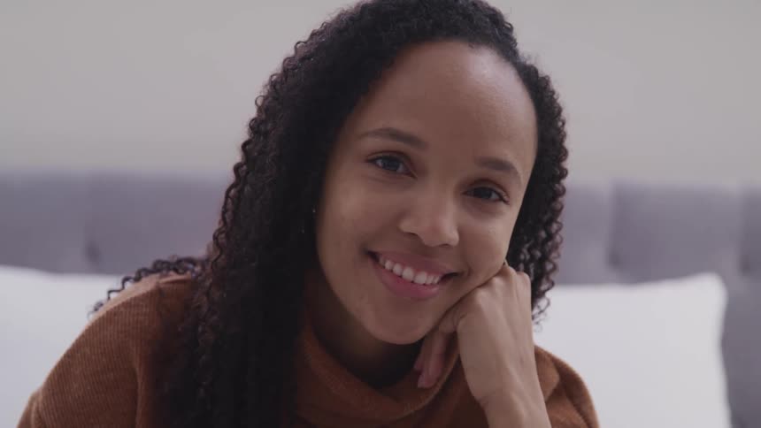 Black woman with curly hair and earrings sitting on a bed and faces the camera with eye contact and smiles broadly