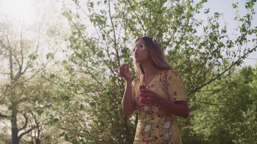 A woman stands outdoors using a bubble wand to bubbles into the air