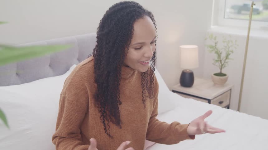 Woman talks and waves on a video call while sitting in bed in a bedroom