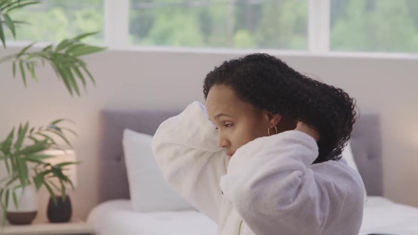 A woman sits on her bed and puts an earring in and fixes her hair