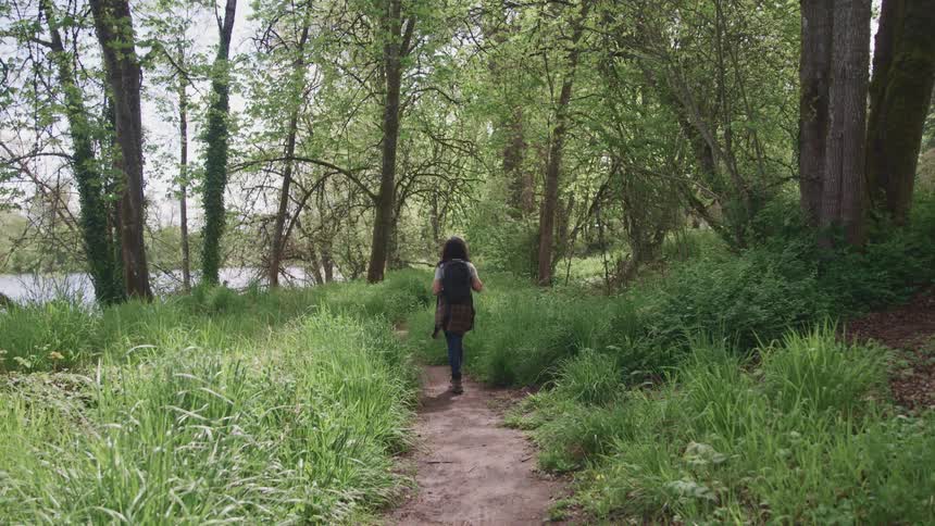 Woman with a black backpack and jeans walks along a dirt track in the forest