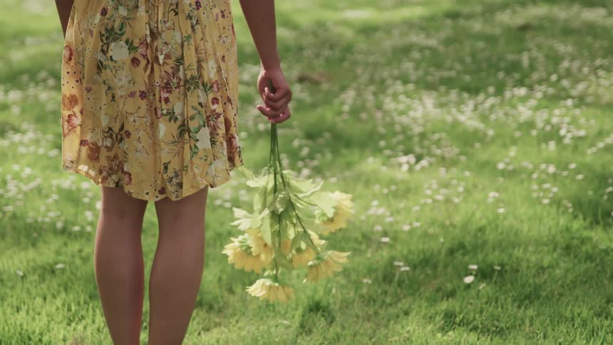 A woman stands in a field wearing a yellow sundress and holding a bouquet of yellow flowers