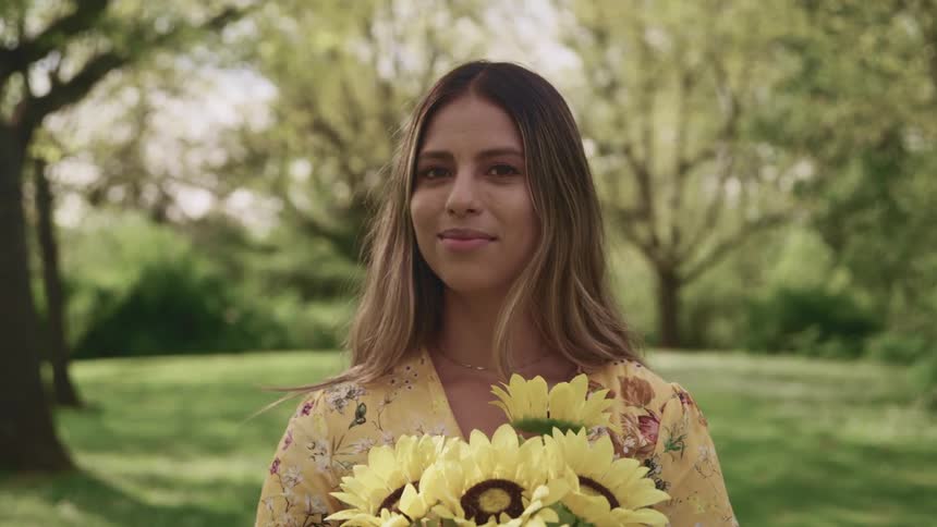 Woman with brown hair a yellow dress and holding a bunch of sunflowers stands on grass and faces the camera with eye contact