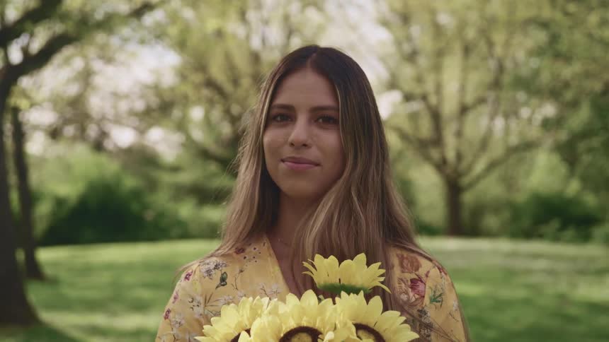 Woman with brown hair yellow dress holds a bunch of sunflowers and faces the camera with eye concat in a public park
