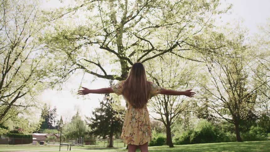A woman faces the sky and slowly raises her arms wide as she stands in a park on a sunny day