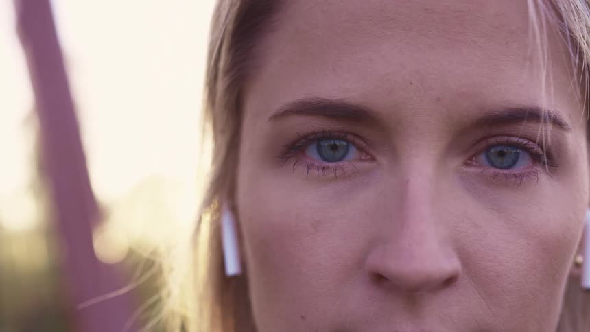 Blonde woman with blue eyes and white wireless earbuds stares at the camera