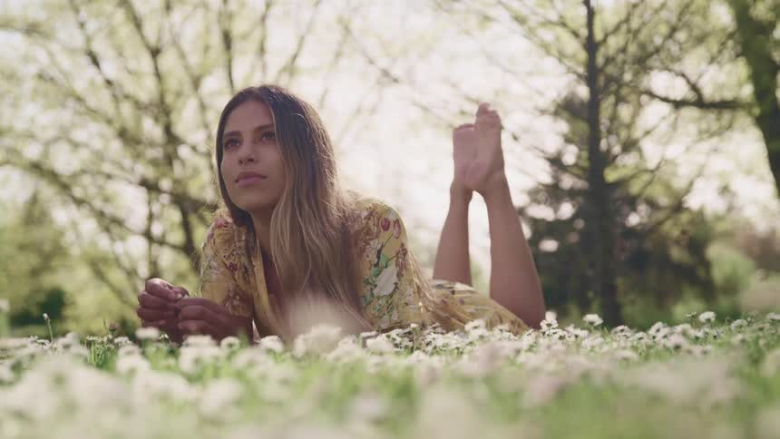 A young barefoot woman lies on her stomach among white flowers in a meadow during the day
