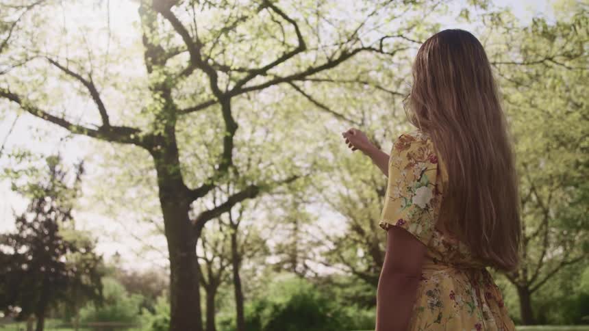 A teenage girl standing in a grassy area with trees raises her arm towards the sky