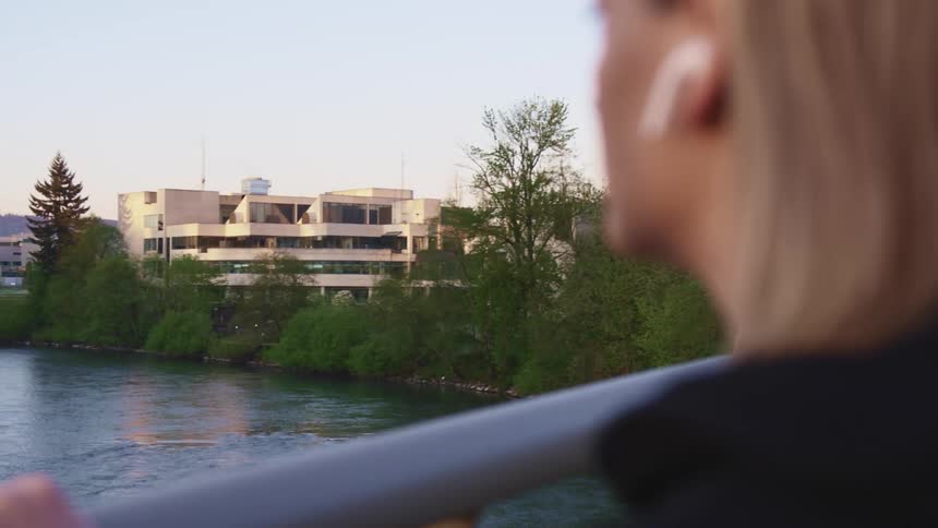 Woman wearing white earbuds reclines on the railing of a bridge looking at a river