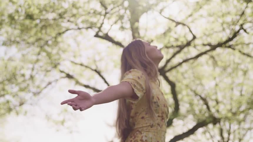 A teenager stands under the trees with her arms spread to her side as she looks up