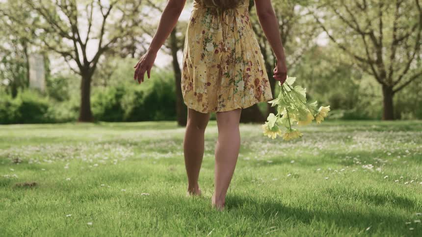 Woman wearing sundress walks barefoot across lawn carrying a bouquet of sunflowers