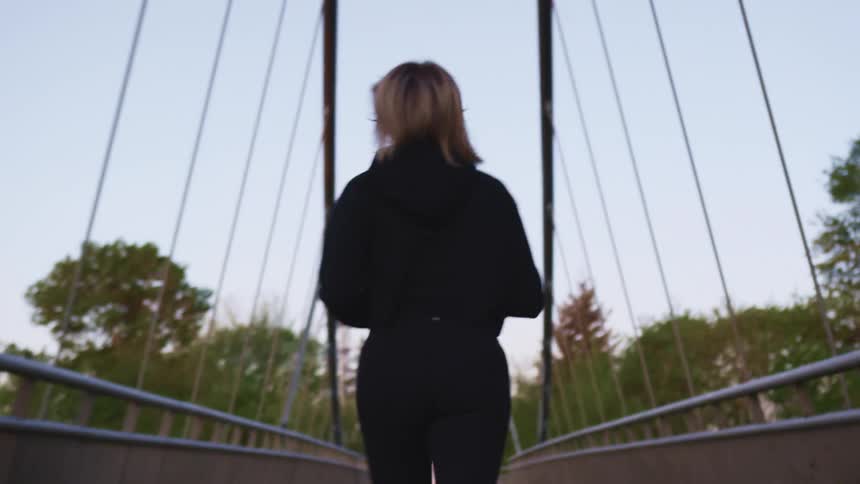 Blonde woman with a black jacket and pants runs across a bridge at dusk