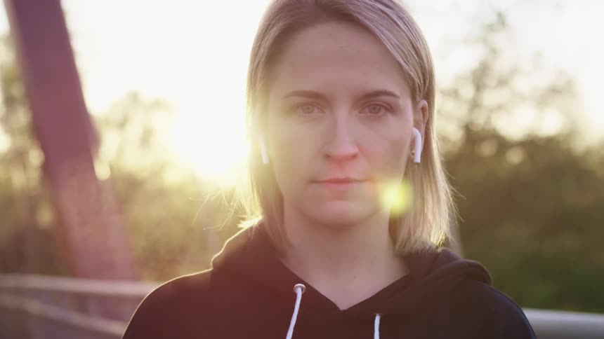 Blonde woman with a black hoodie and white wireless earbuds stares at the camera as the sun shines in the background brightly