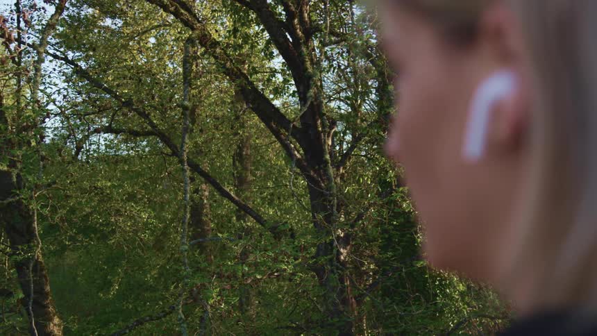 Close up of a womans face wearing white earbuds while looking at the trees
