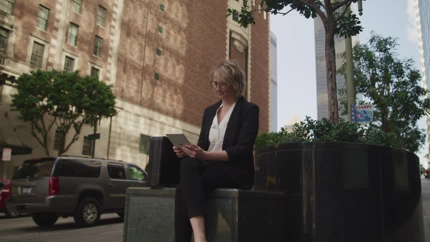 A woman sits on a box on the sidewalk outside a city building reading a tablet and dressed as a business professional