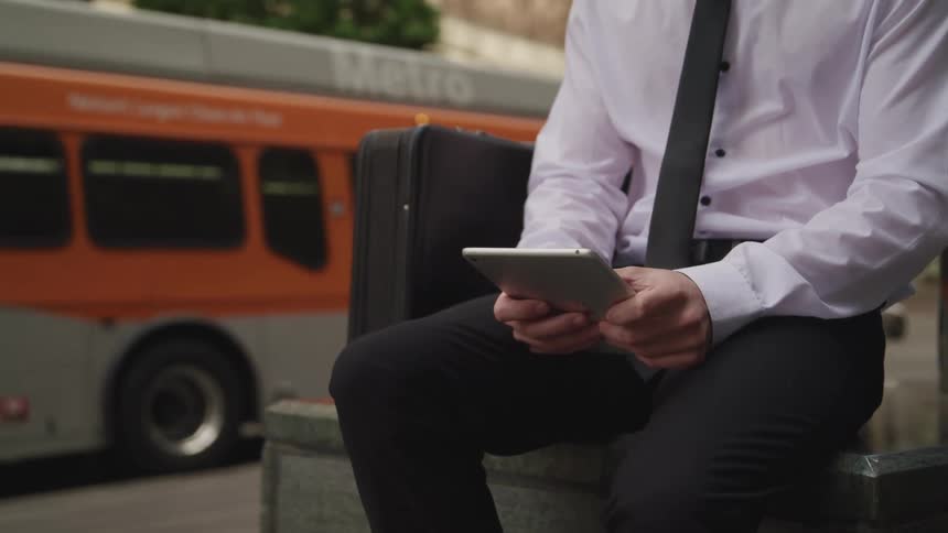 A man sits outside and uses a tablet as a bus drives past