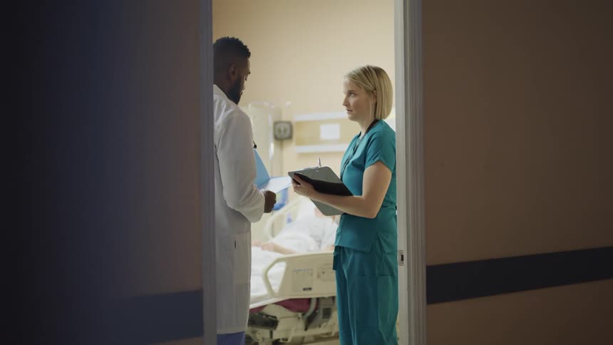 Nurse and doctor stand in hospital room next to patient while she takes notes on clipboard and listens to doctor