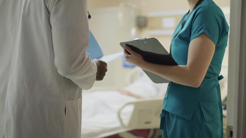 A doctor and nurse talk in a hospital room while a patient lies in the bed