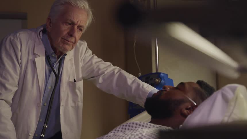 An elderly doctor stands at bedside looking at a sleeping patient with concern